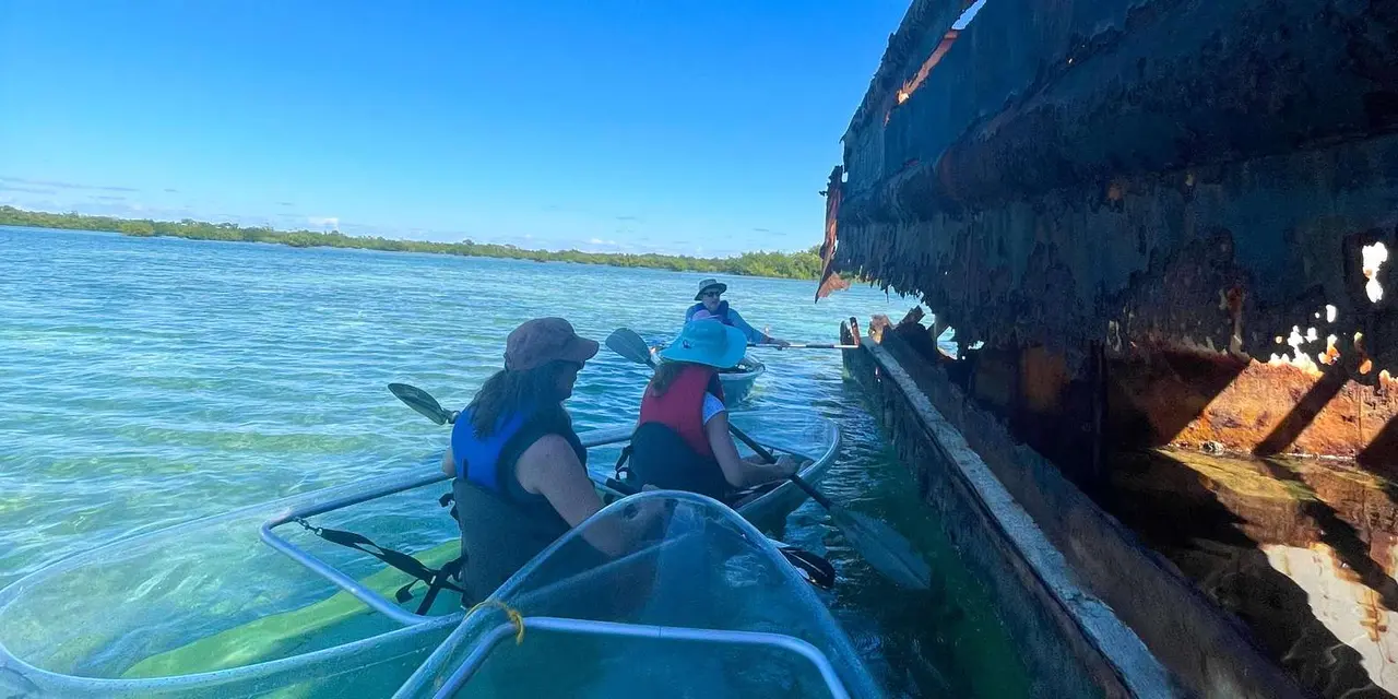 Paddlers in clear kayaks on calm turquoise water alongside the rusted hull of a wreck