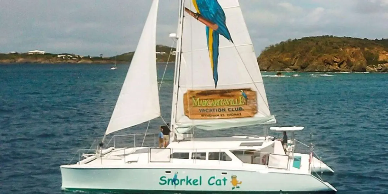 The white Snorkel Cat catamaran under sail on dark blue water, a parrot painted on its sail and green islands behind