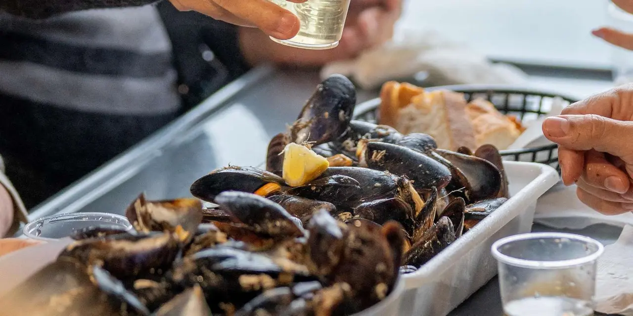 A hand pouring white wine into a glass beside a tray of steamed mussels on a boat table