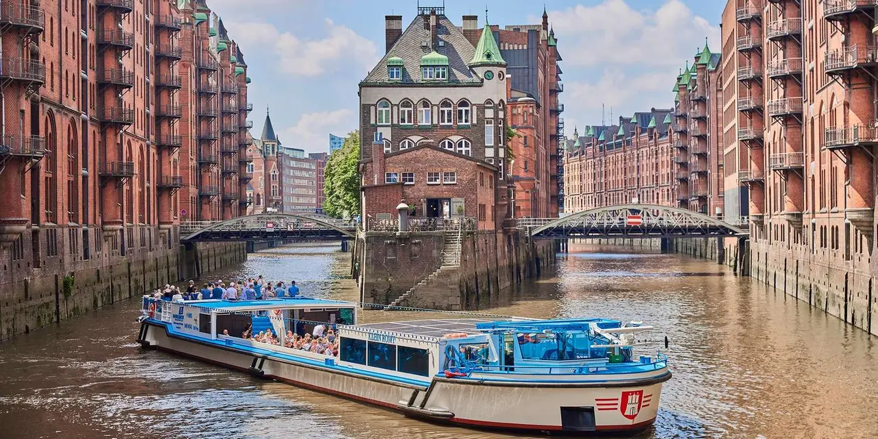 A white-and-blue harbor tour boat full of passengers gliding down a Speicherstadt canal between red-brick warehouses, with the Wasserschloss building and iron bridges ahead