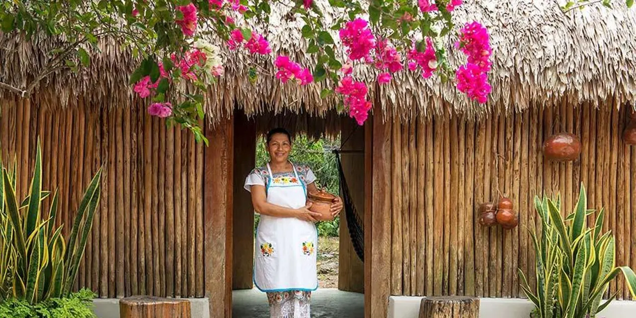A woman in an embroidered white apron holding a clay pot in the doorway of a thatched wooden hut under pink bougainvillea