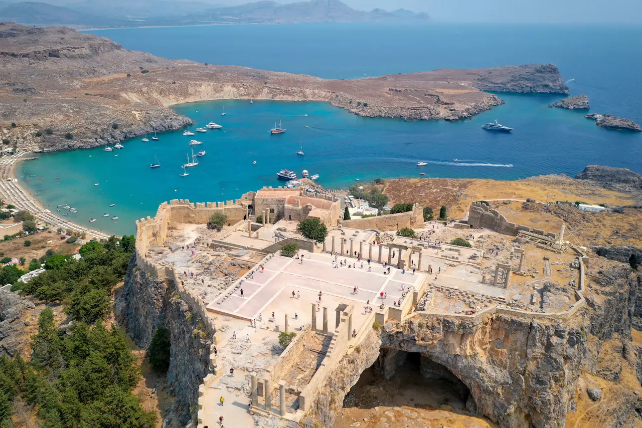 Aerial view of the Acropolis of Lindos, with its walls and columns on a rocky hilltop above a turquoise bay of moored sailboats and a long beach; Lindos is about 50 km from Rhodes Town