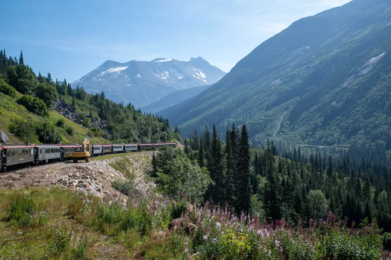 A passenger train curving along a green mountainside above a forested valley, with snow-dusted peaks and blue sky beyond, near Skagway, Alaska