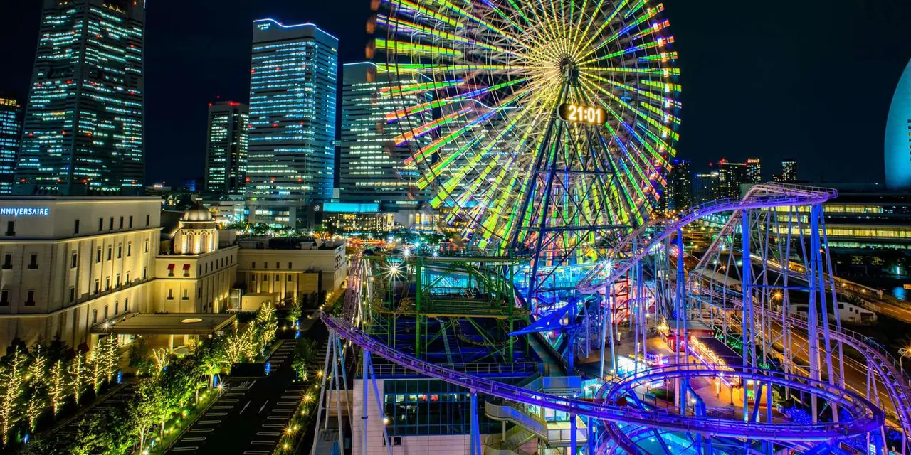Yokohama's Cosmo Clock 21 ferris wheel lit in green, yellow and blue at night beside a roller coaster, with Minato Mirai office towers behind