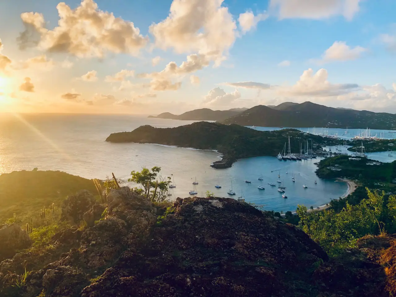 Golden-hour sun over a wide bay on Antigua, with sailboats at anchor and green hills across the water