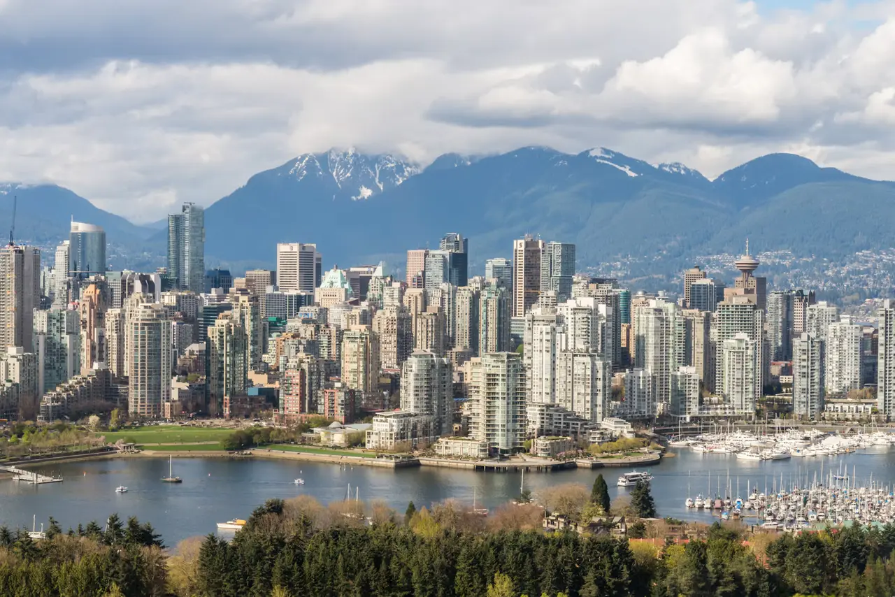 Downtown Vancouver's high-rises with the Harbour Centre tower, snow-dusted North Shore mountains behind and a marina of sailboats on the water below, framed by evergreens under a cloudy sky