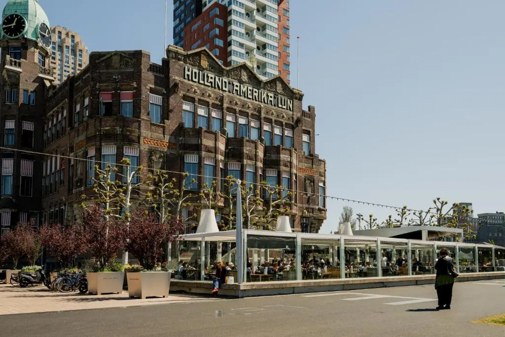 Hotel New York's brick 1901 building with the Holland Amerika Lijn lettering across the facade and a glass-walled terrace restaurant in front