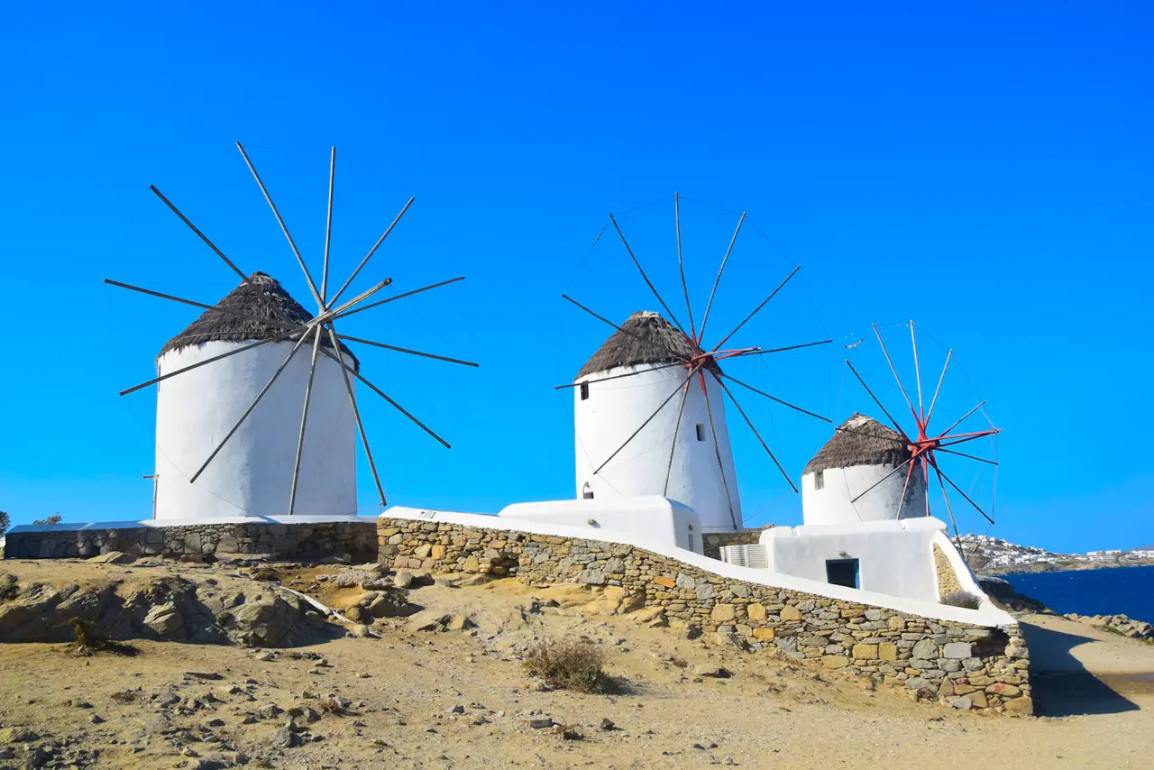 Three white windmills with thatched roofs and wooden sails on a stone terrace above Mykonos Town, under a deep blue sky