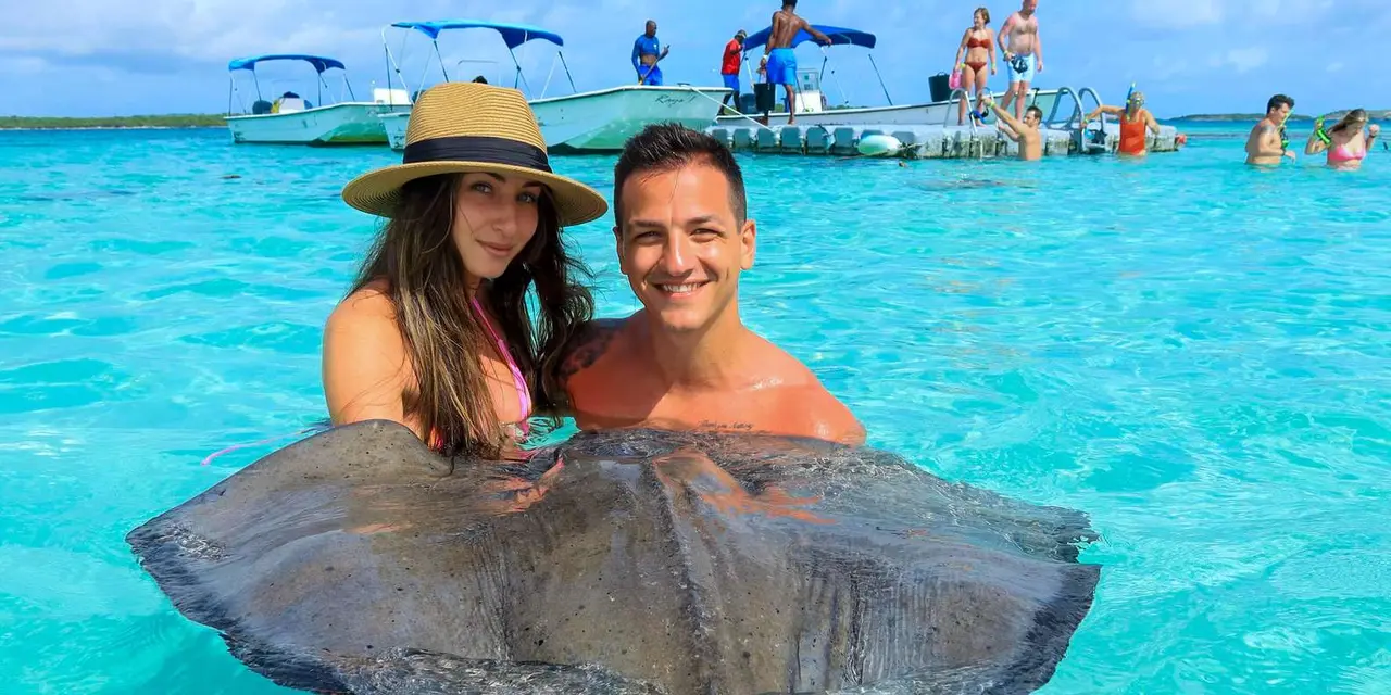 A smiling couple standing in turquoise shallows holding a large stingray, with boats and a floating platform behind them