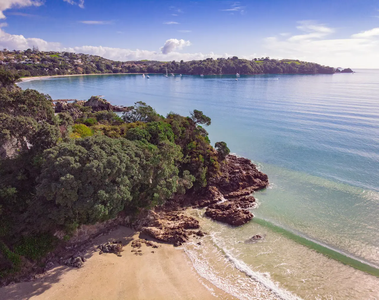 Aerial view of a sandy cove on Waiheke Island, with pōhutukawa-covered rocks, clear green water and yachts moored in the bay — about 40 minutes by ferry from Auckland's cruise wharves