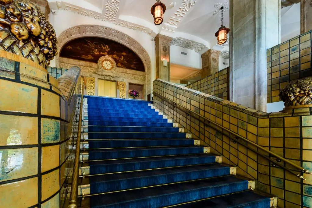 Blue-carpeted grand staircase in the lobby of Hotel New Grand, Yokohama, with tiled balustrades, gilded fixtures and a clock above the landing