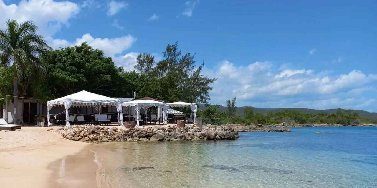A curved white-sand beach with clear shallow water and white tents under palm trees