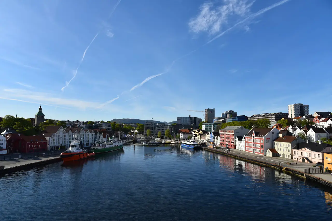 Stavanger's Vågen harbour on a sunny morning, a red tug and a green ship moored beside rows of white and pink timber houses, with hills and a clear blue sky behind