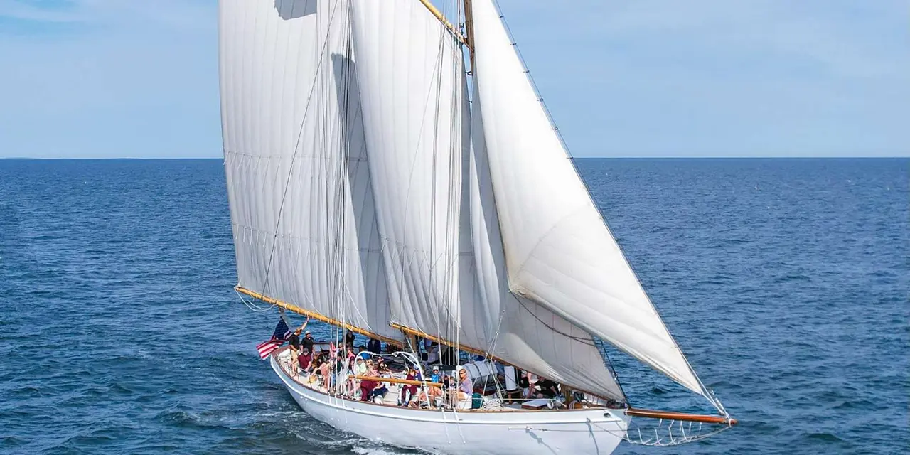 A white schooner under full sail with passengers aboard on open blue water