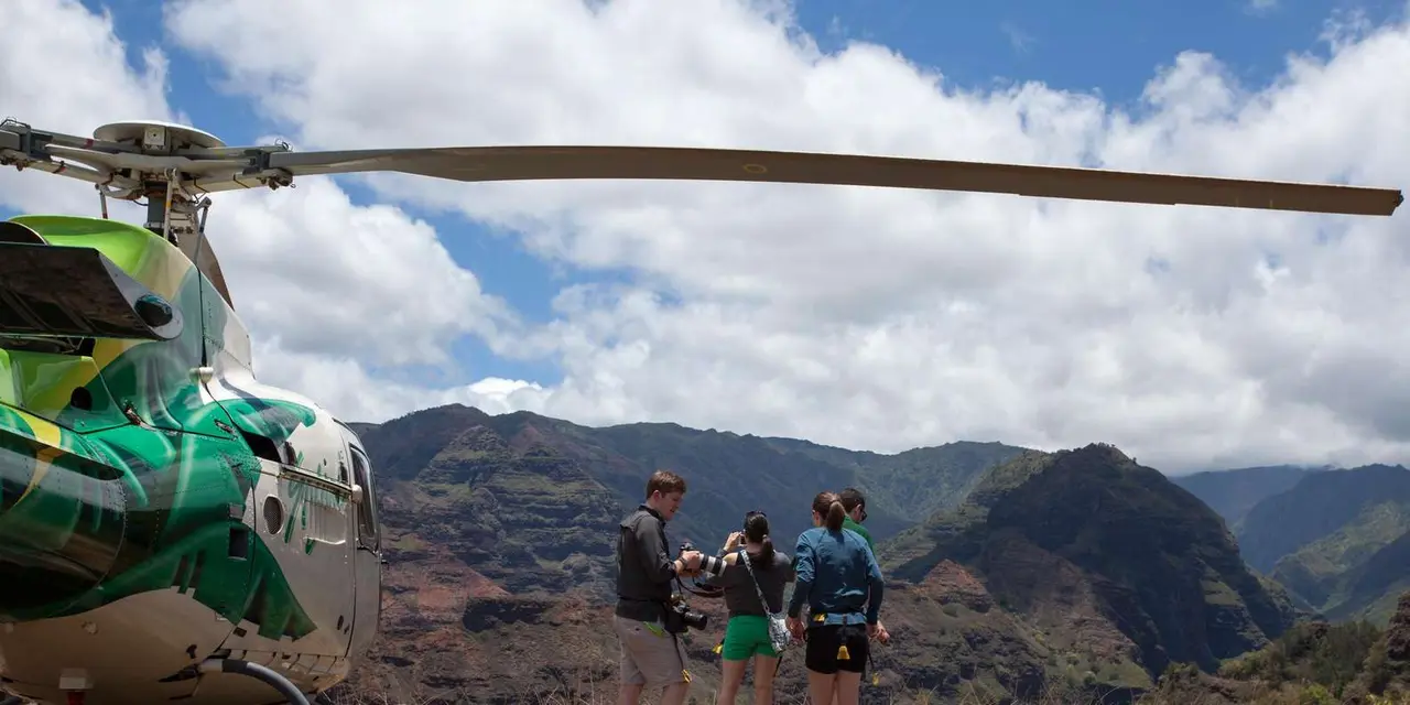 A green-and-white helicopter parked on a landing pad while passengers look out over a canyon under a cloudy sky