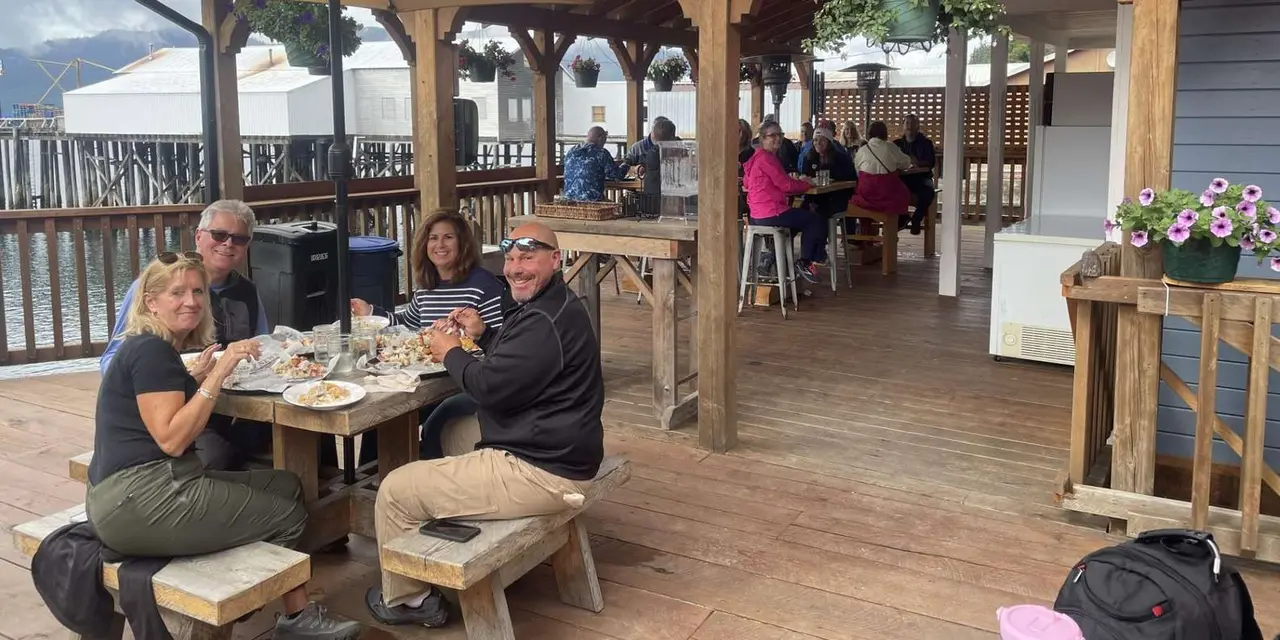 Four guests smiling over plates of crab at wooden tables on a covered deck above the water, with old cannery buildings behind