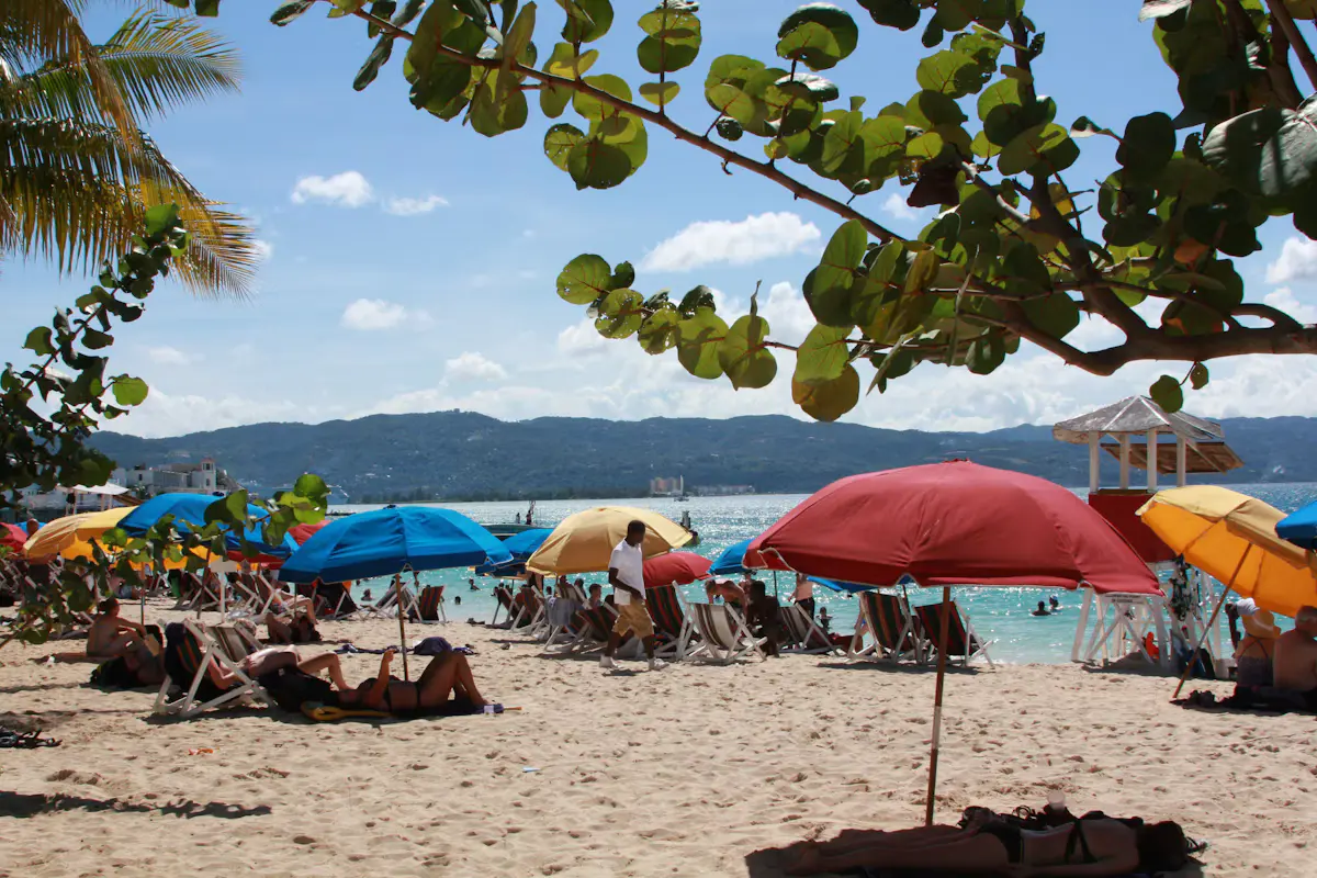 Red, blue and yellow umbrellas on a sandy beach at Montego Bay, Jamaica, west along the coast from Falmouth, with palm branches overhead and hills across the bay