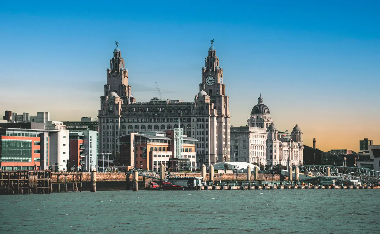 The Royal Liver Building and the Port of Liverpool Building across the River Mersey at sunset, the twin clock towers and domes glowing against a deep blue sky fading to orange