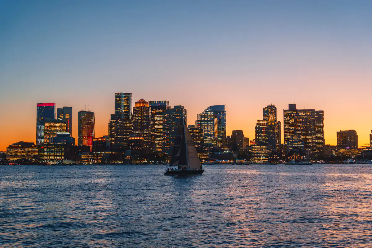 A sailboat crosses Boston Harbor at sunset, the downtown Boston skyline lit up against an orange and blue sky