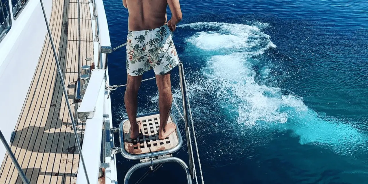 A man standing on a boat's swim platform above deep blue water, with swimmers jumping off the deck behind him