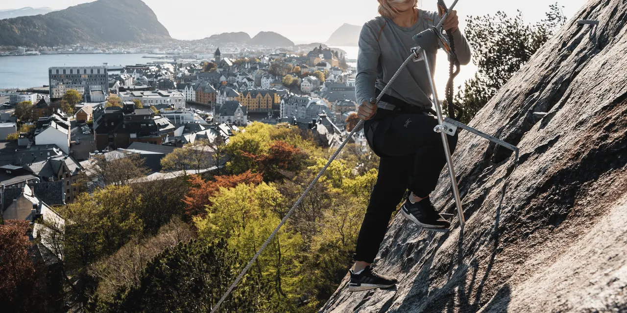 A climber in a helmet clipped to a cable on a rock face, with Ålesund's rooftops and the sea far below