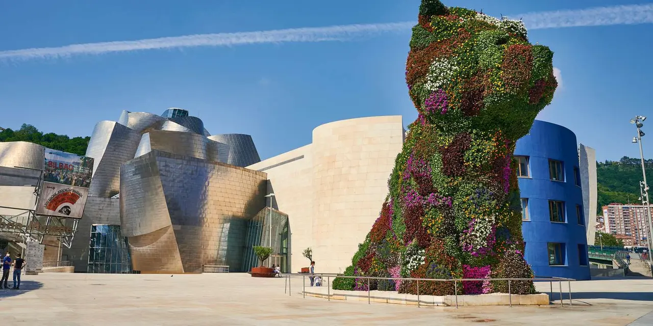 A giant flower-covered dog sculpture beside the Guggenheim's metal curves under a blue sky