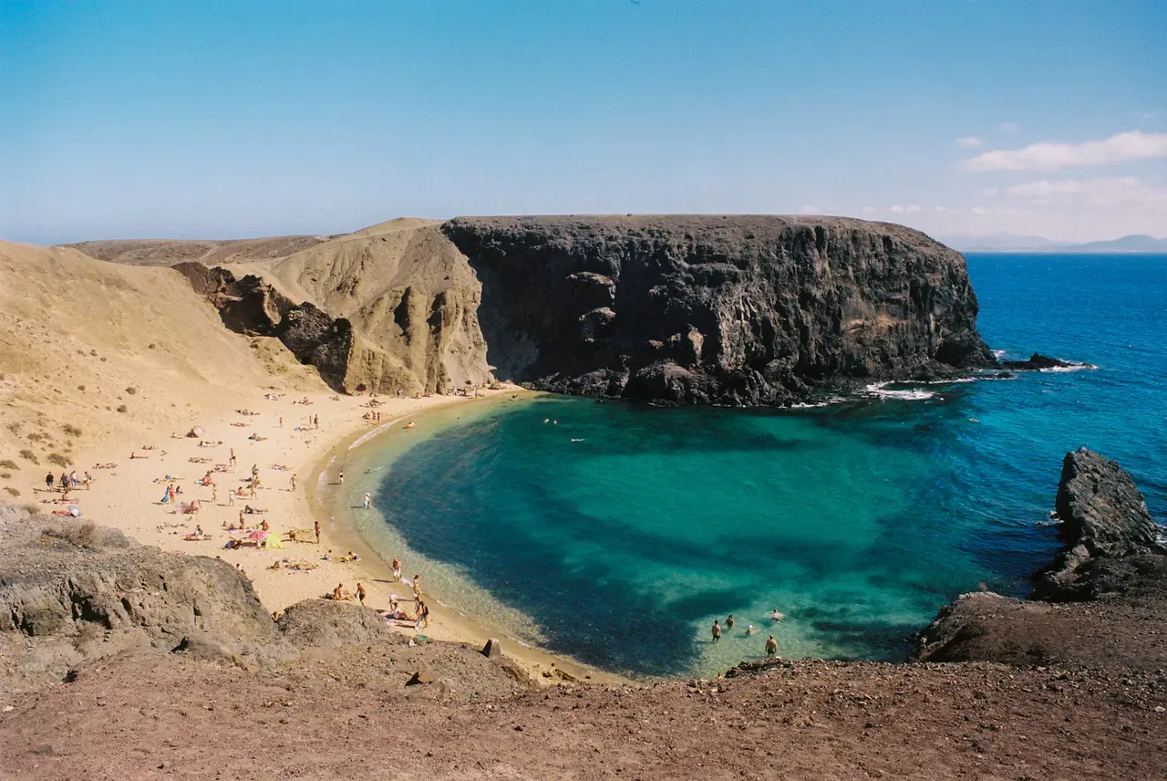 Turquoise water in the sheltered cove of Playa de Papagayo below dark cliffs, with sunbathers on the sand, on Lanzarote's southern tip about 18 miles from the cruise port