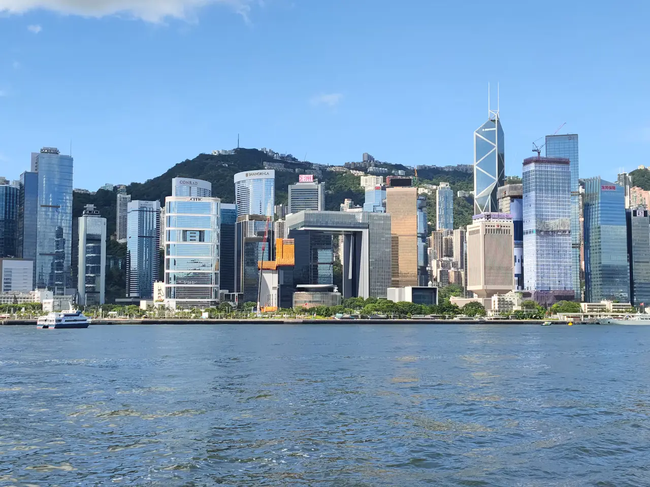 Hong Kong Island's skyline of glass towers along Victoria Harbour on a clear day, with green hills behind and blue water in front