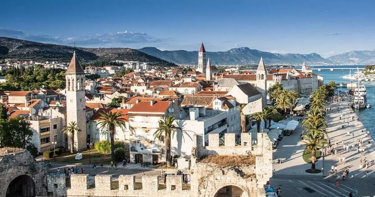 Trogir's red roofs, bell towers and palm-lined waterfront seen from the fortress walls, with mountains behind