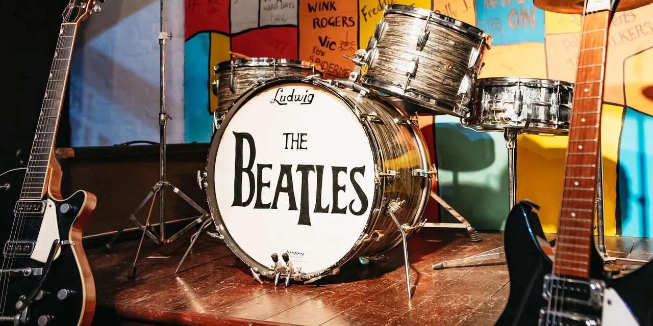 A drum kit with 'The Beatles' painted on the bass drum, flanked by two guitars on a small stage in front of a hand-painted backdrop