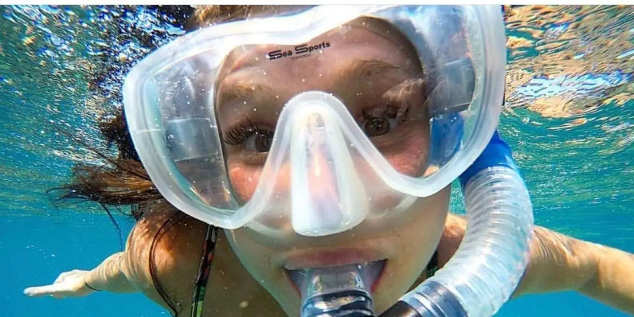 A snorkeler in a clear mask and snorkel looking into the camera just under the surface of bright blue water