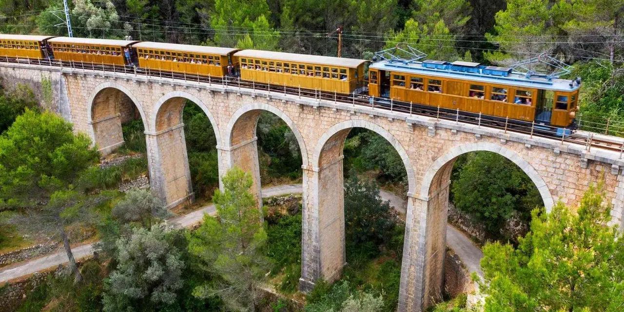 A yellow wooden train crossing a stone arch viaduct among pine trees in the Tramuntana mountains