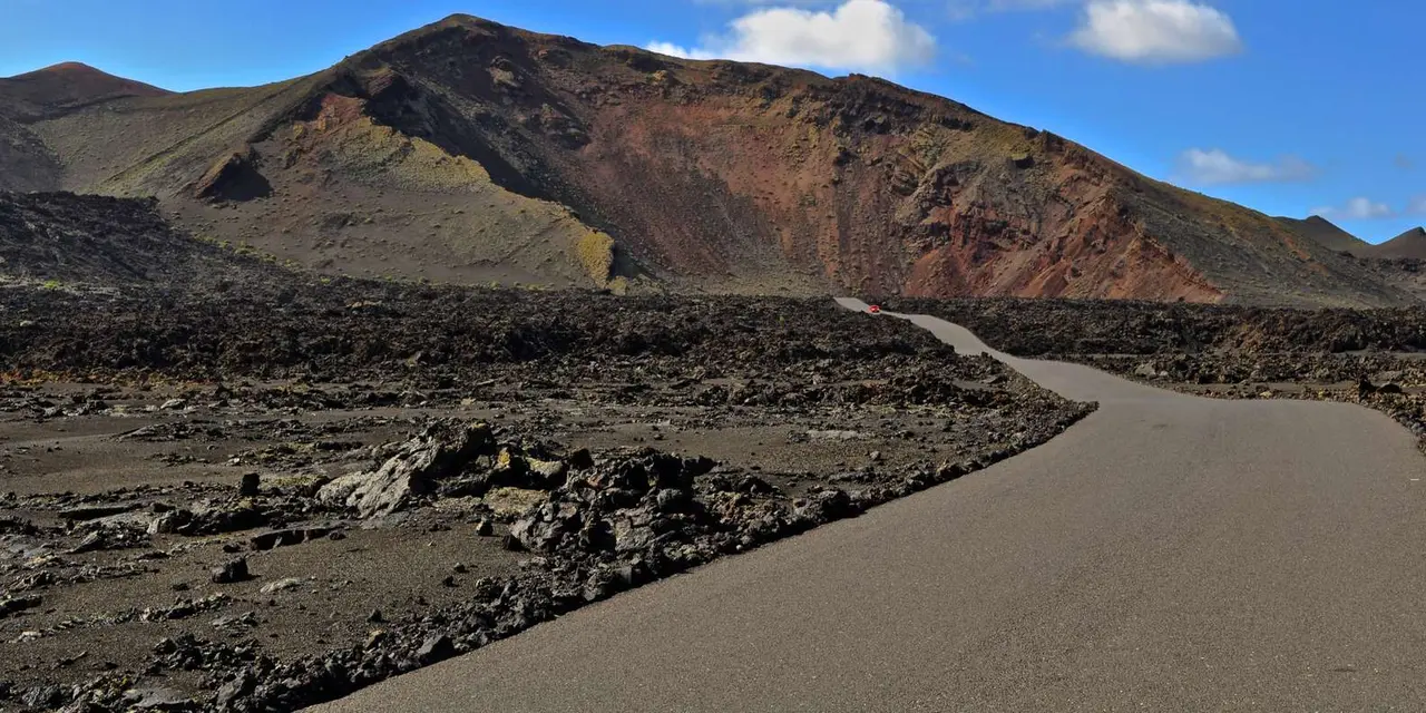 A road curving through a black lava field below a red-brown volcano under a blue sky
