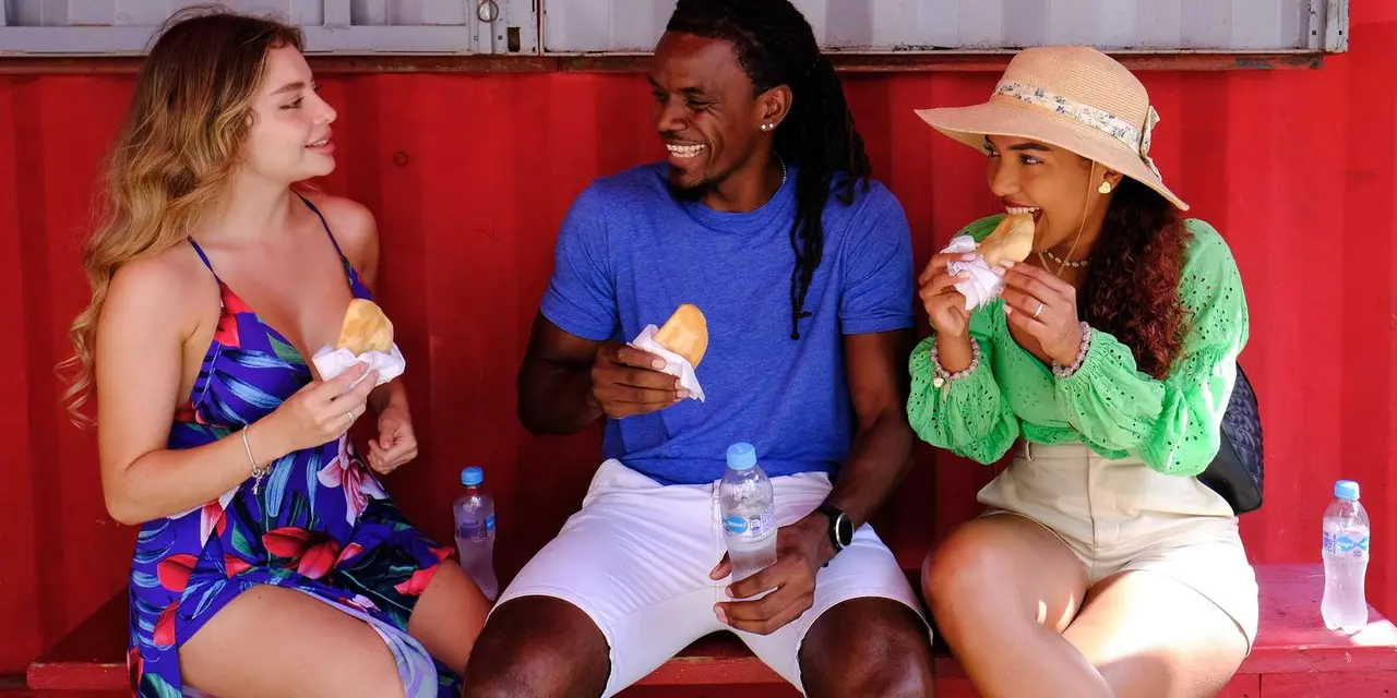 Three smiling people sitting on a red bench against a red wall, eating golden fried pastries with water bottles beside them