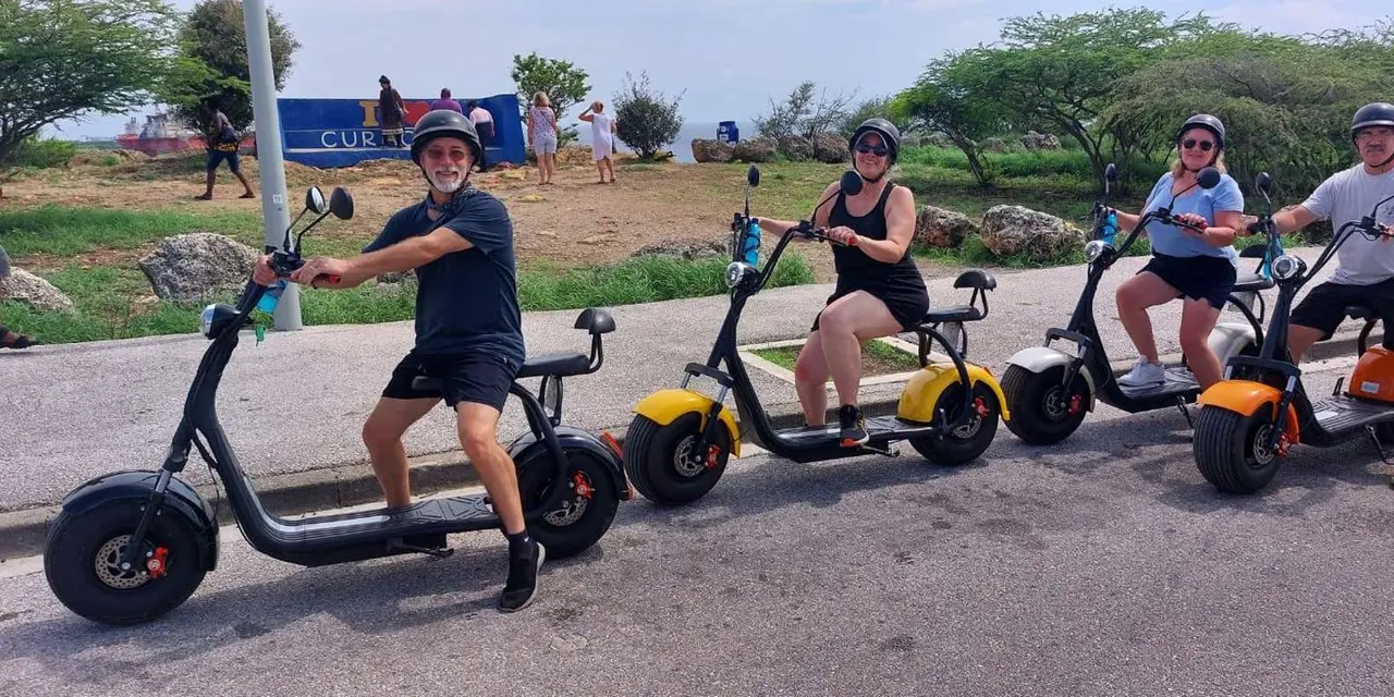 Smiling riders in helmets on fat-tyre electric scooters on a road lined with dry scrub