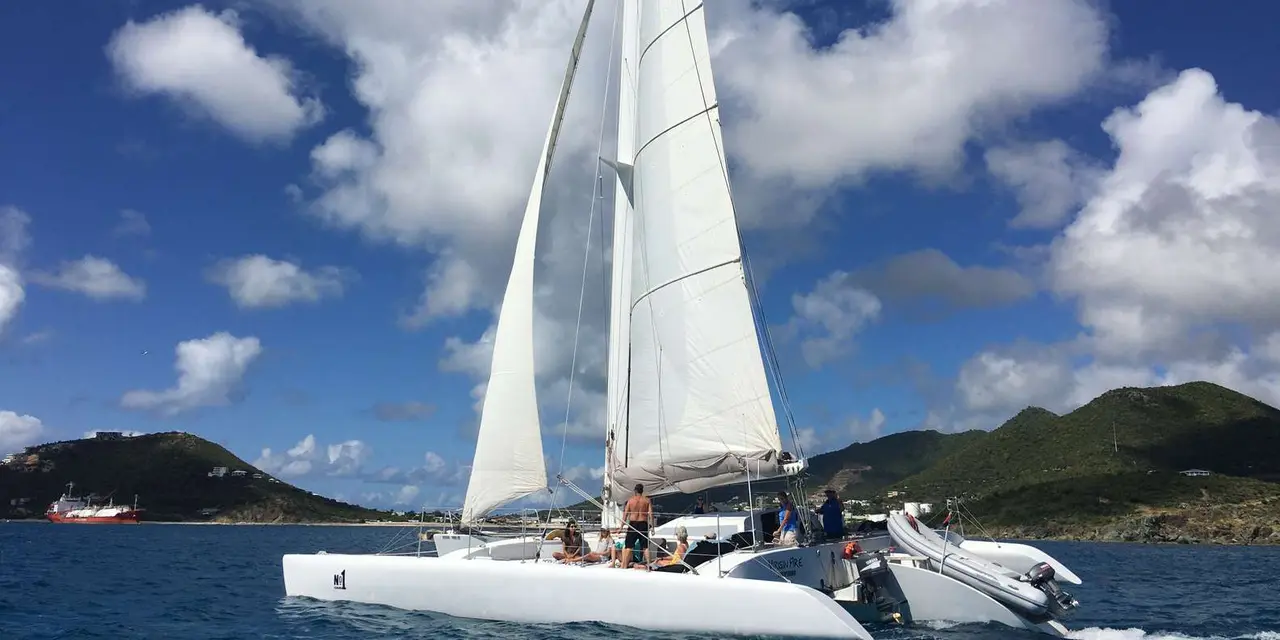 A white trimaran under sail in blue water, with green hills behind