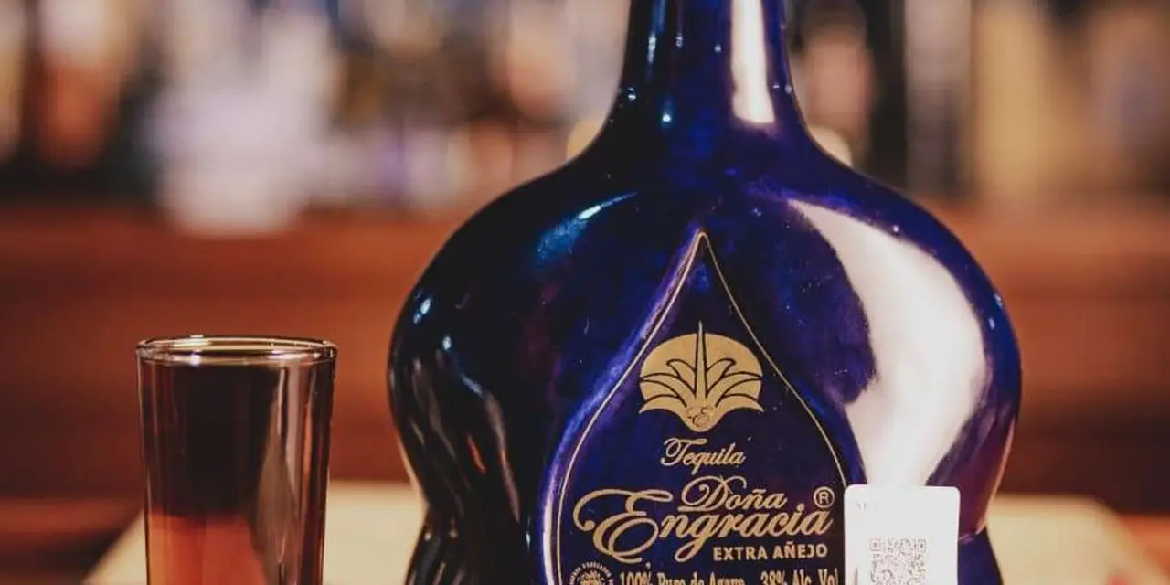A cobalt-blue Dona Engracia tequila bottle beside a shot glass of amber tequila on a bar with a blurred backdrop
