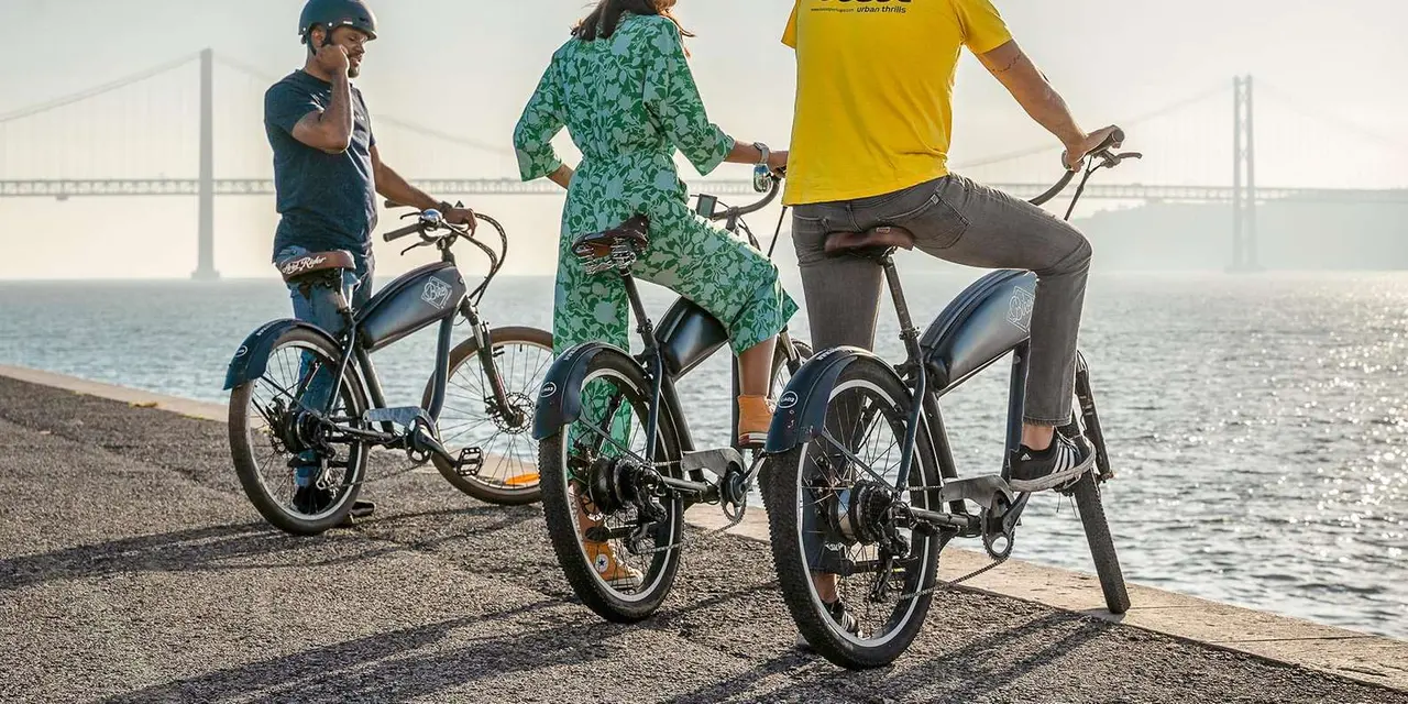 Three cyclists on e-bikes beside the Tagus with the 25 de Abril Bridge hazy in the background