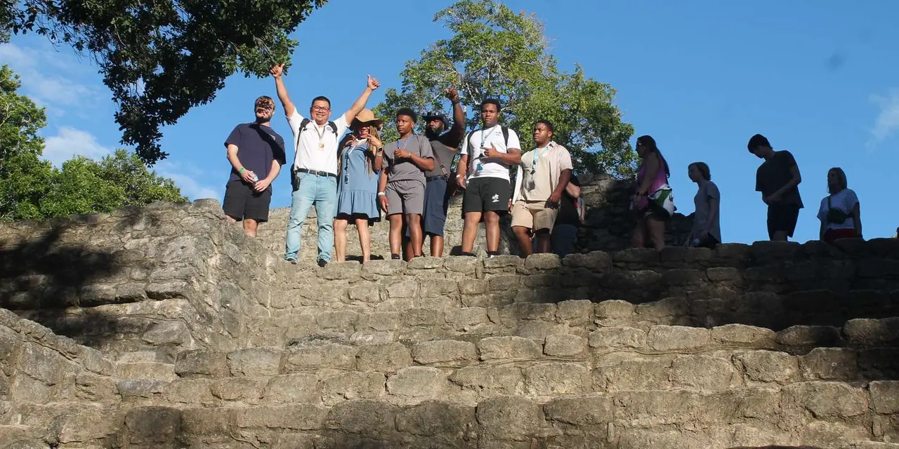 A guide with raised arms and a group of visitors at the top of a worn stone temple staircase at Chacchoben, with jungle trees and blue sky behind them