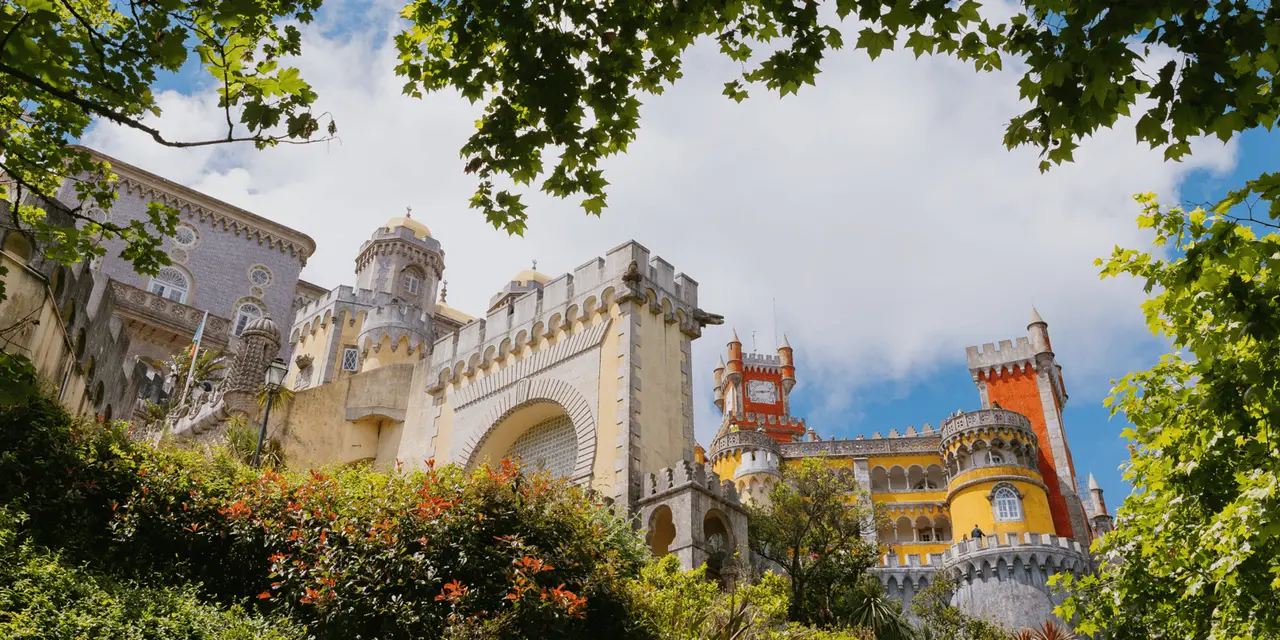 The yellow and red towers and crenellated walls of Pena Palace seen from below through green leaves