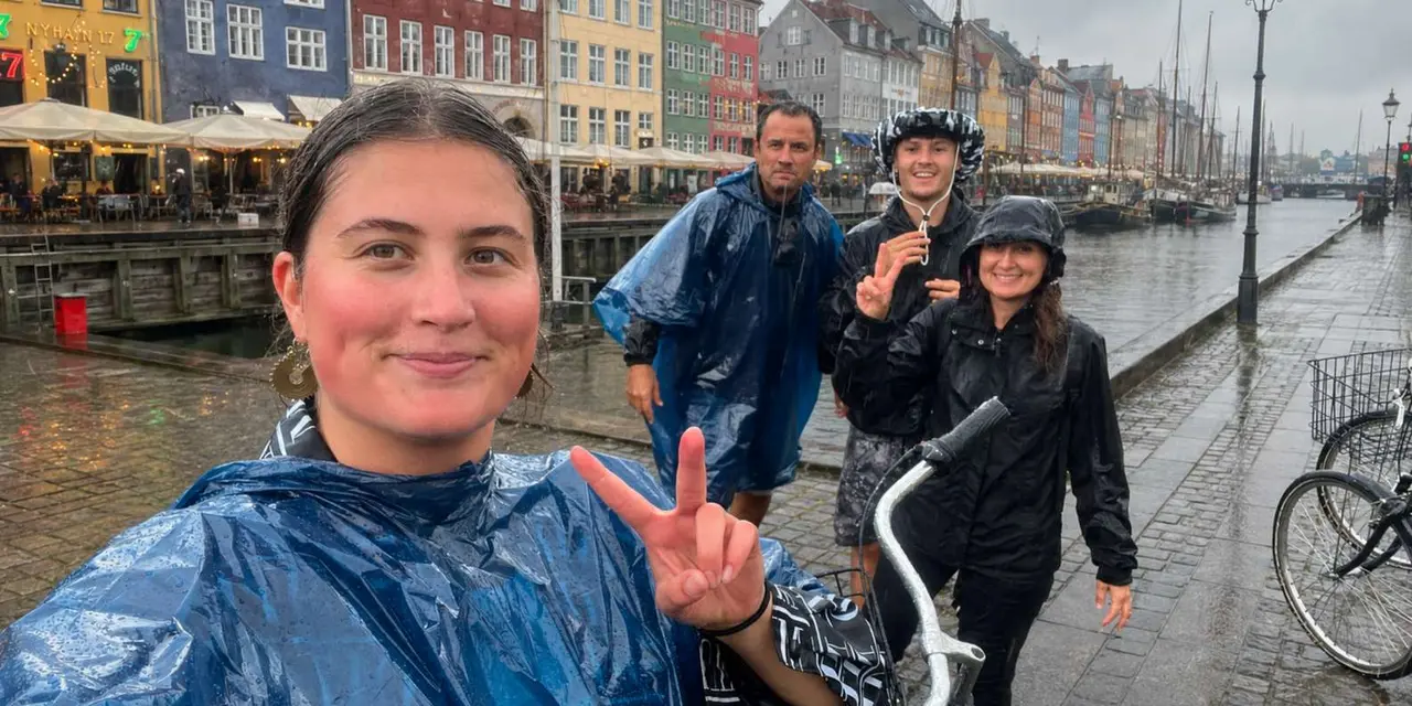 Four people in rain ponchos with a bike on the wet cobbles of Nyhavn, with the colorful canal-side houses and moored sailing ships behind them