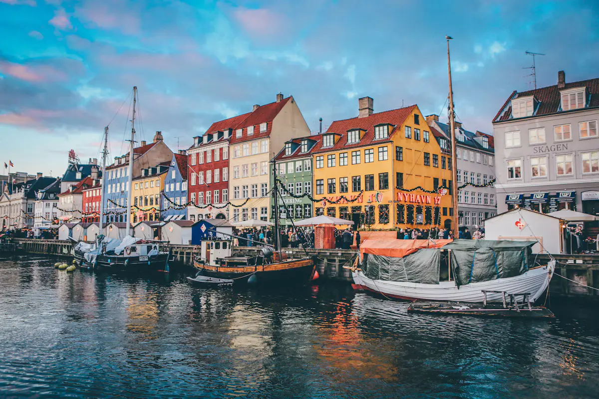 Nyhavn at dusk, with red, yellow, blue and green townhouses strung with lights above wooden boats moored along the canal under a pink and blue sky