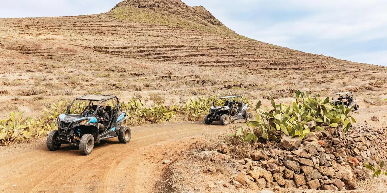 Two off-road buggies on a dirt track among prickly-pear cactus below a dry brown hill
