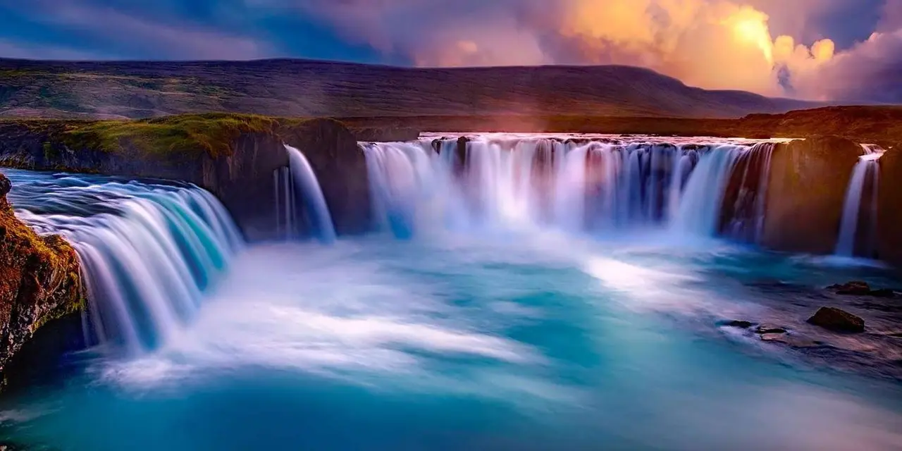 Long-exposure photo of Goðafoss, a wide curved waterfall pouring into a turquoise pool under a glowing sunset sky
