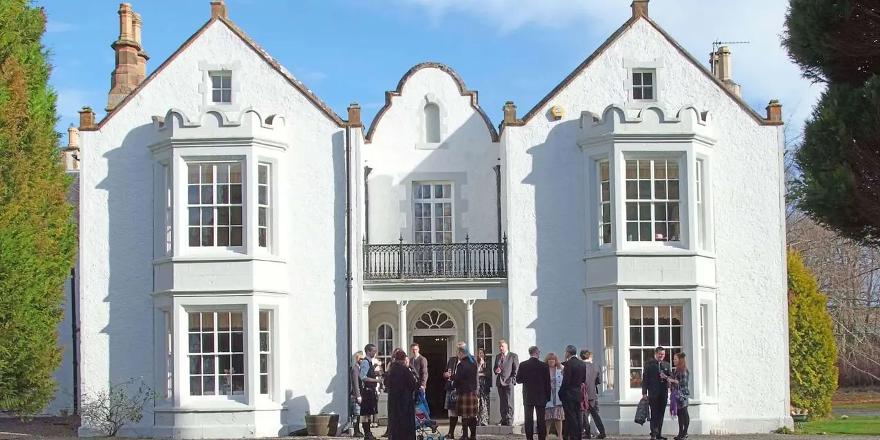 A white Georgian country house with bay windows and a balcony, guests gathered on the gravel in front