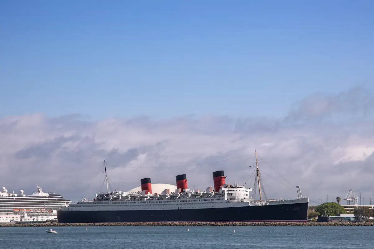 The Queen Mary's red and black funnels above Long Beach harbor under a blue sky, with a cruise ship docked at the left edge