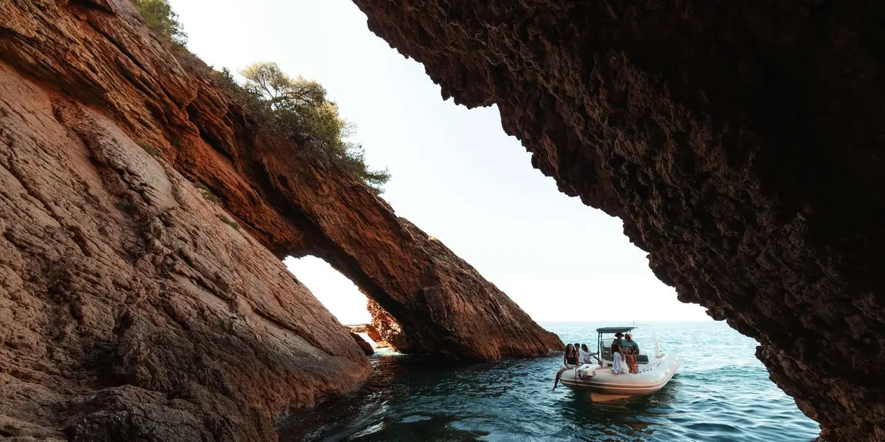 A small boat with passengers passing beneath a red-rock sea arch on clear green water