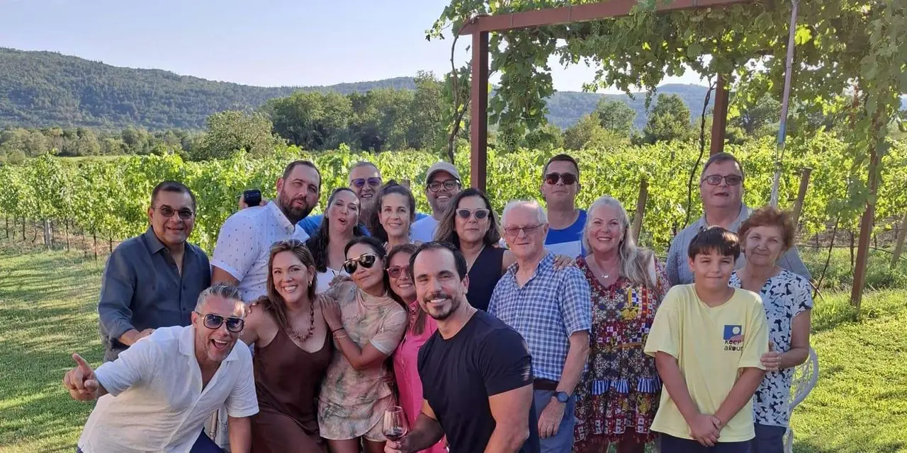 A large group of smiling visitors standing under a vine-covered pergola at a vineyard