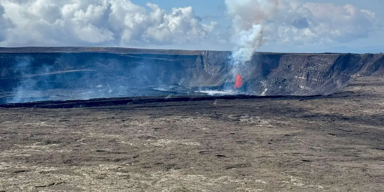 A wide volcanic crater with steam rising and a small fountain of lava on its far side