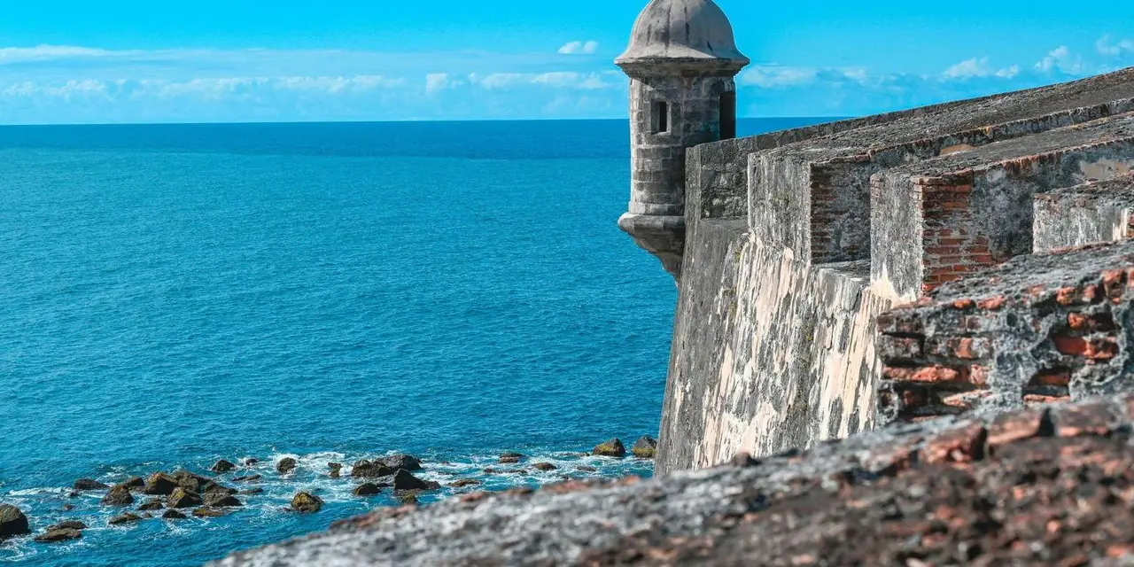 A sandstone sentry tower on a fort wall beside the sea under a blue sky